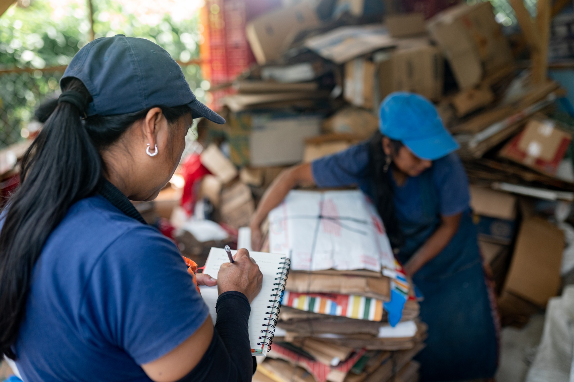 Hispanic Woman Noting Recycled Cardboard Inventory While Teammate Handles Stacks in Warehouse.