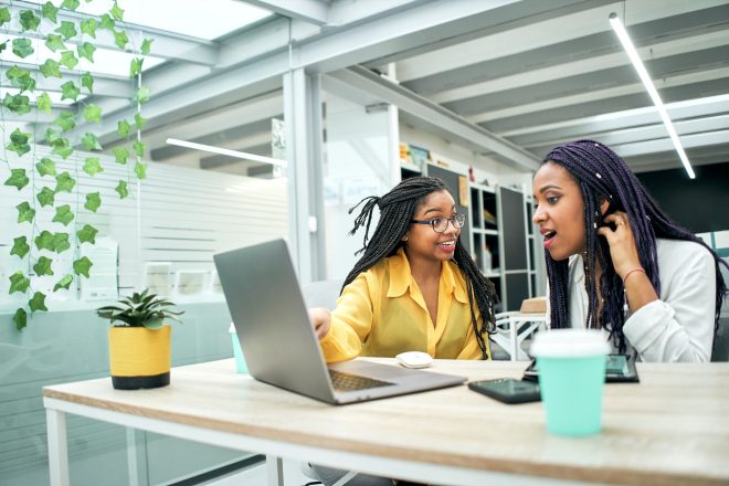 Two young women in office looking surprised at something they are seeing on laptop screen.