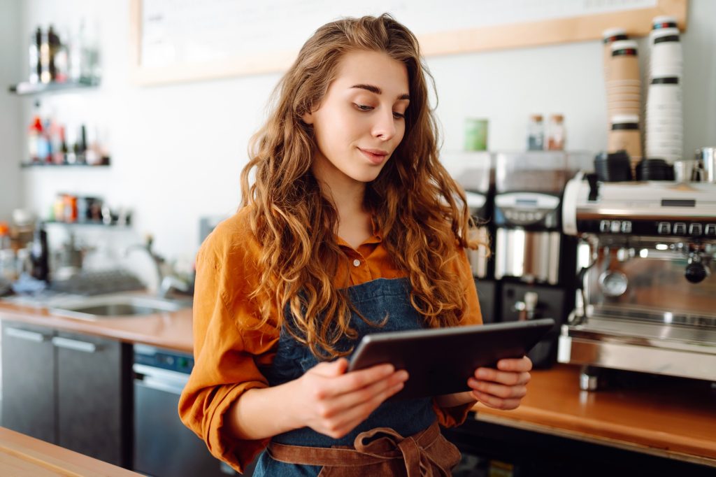Young woman in apron with digital tablet at bar counter of coffee shop takes order. Small business.