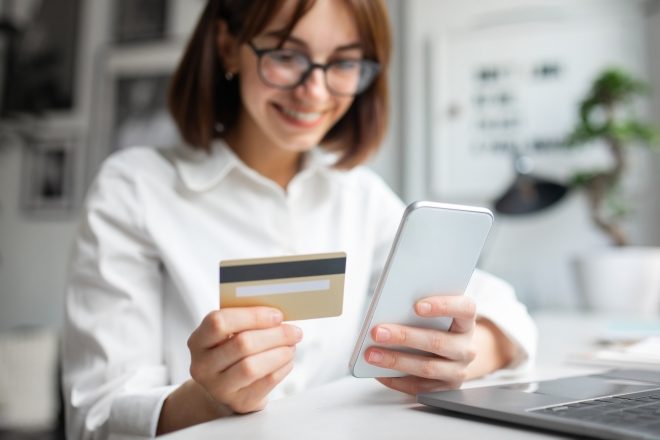 Young woman using smartphone and credit card, sitting at table with laptop, using modern mobile app